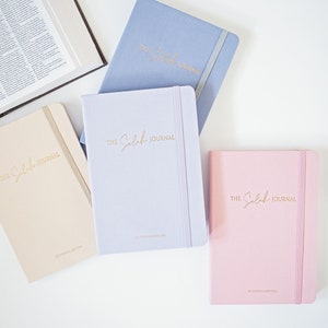 May include: Four journals in pastel colors: beige, lavender, light blue, and pink. Each journal has the gold text "The Seleh Journal" and a matching elastic band. An open book is in the background.