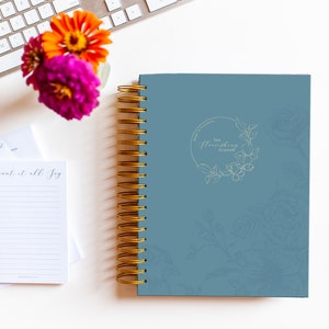 Peut inclure: Un agenda spirale bleu avec des accents dorés et un motif floral. La couverture indique "The Flourishing Planner".