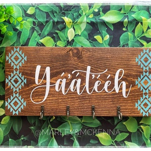 May include: A wooden key holder with the text "Yá'át'éeh" in white lettering. The key holder has four hooks and is decorated with a turquoise and white geometric pattern on the sides. The key holder is sitting on a green leafy background.