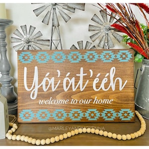 May include: Wooden sign with turquoise and white geometric patterns, the text "Yó'át'ééh welcome to our home" and the social media handle "@MARLEYANDMCKENNA".