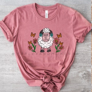 May include: A pink t-shirt with a white sheep wearing headphones and surrounded by colorful flowers and butterflies. The text "@MARLEYANDMCKENNA" is printed on the bottom of the shirt.