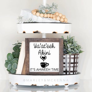 May include: A two-tiered white tray with a rustic finish, decorated with greenery, wooden beads, and a framed sign that reads "Ya'at'eeh Abini It's Ahwee'eh Time".