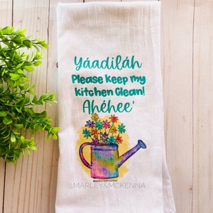 May include: White kitchen towel with a colorful watering can and flowers. The text "Yaadiláh Please keep my kitchen Clean! Ahéhee'" is printed on the towel. The text "@MARLEY&MCKENNA" is printed at the bottom of the towel.