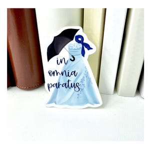 May include: A sticker featuring a light blue dress with a black umbrella and a blue bow. The sticker has the Latin phrase "in omnia paratus" in a handwritten font. The dress has floral embroidery.