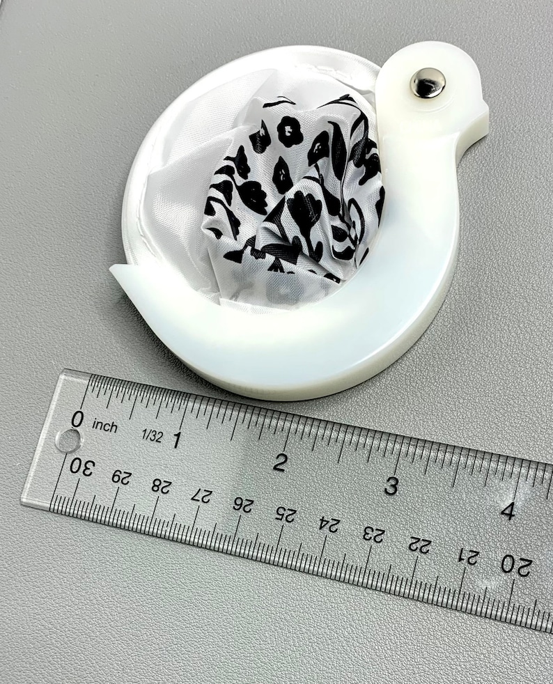 Op de afbeelding: Een witte, zwaanvormige thee-infuser met een theezakje met zwart-wit bloemenpatroon erin. De infuser is ongeveer 7,6 cm lang. Een transparante liniaal is naast de infuser geplaatst voor de schaal.