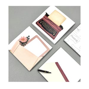 May include: A selection of square Post-it notes with different designs. One shows a red typewriter, another a pink envelope with a flower, and a third with a blank cream-coloured paper. A black pen rests on a white paper with a red border.