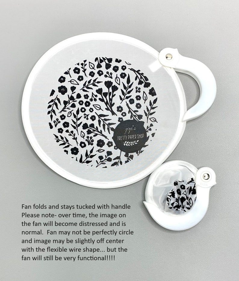 Op de afbeelding: Witte, ronde handventilator met een handvat, met een zwart bloemenontwerp. De ventilator bevat de tekst "jojo's PRETTY PAPER SHOP". De ventilator vouwt op en blijft opgeborgen met het handvat.