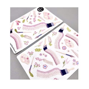 May include: Two white sticker sheets with illustrations of art supplies. The stickers feature paintbrushes with blue handles, paint palettes, paint tubes, and floral accents in shades of pink, purple, and green. The text "original hand drawn art" is visible on one sheet.
