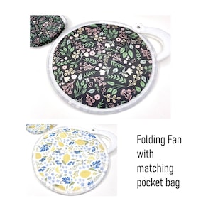 May include: Folding fan with a matching pocket bag. The fan has a white handle and a black background with a floral pattern in pink, white, and green. The pocket bag has the same floral pattern.
