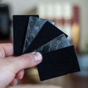 May include: A collection of black VELCRO brand hook and loop fasteners. The fasteners are rectangular with a textured surface. The adhesive backing is visible on some pieces. The word "VELCRO" is printed on the adhesive backing.