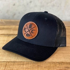 May include: Black and brown trucker hat with a leather patch featuring a crown, crossed swords, and the letters "KC".