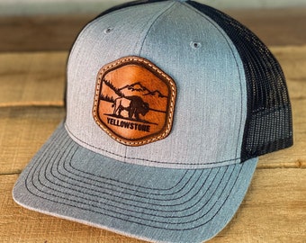 May include: A gray and black trucker hat with a brown leather patch featuring a silhouette of a bison and the text "Yellowstone".