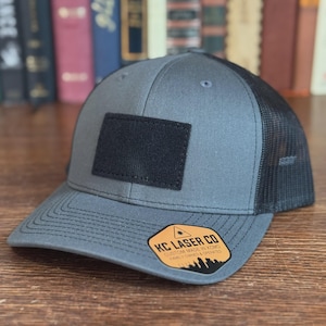 May include: A gray trucker hat with a black mesh back and a black rectangular patch on the front. The hat has a curved brim and a tag that reads "KC LASER CO CUSTOM MADE IN KSMO FAMILY OWNED & OPERATED".