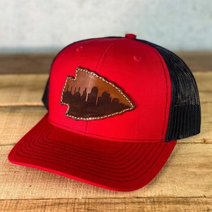 May include: A red trucker hat with a black mesh back and a curved brim. The hat features a leather patch in the shape of an arrowhead, showcasing a city skyline in brown, stitched with white thread.