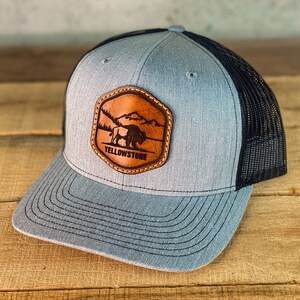 May include: A gray and black trucker hat with a brown leather patch featuring a silhouette of a bison and the text "Yellowstone".
