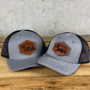 May include: Two gray trucker hats with black mesh backs. Each hat features a brown leather patch with a bear silhouette inside a brown hexagon. The hats are displayed on a wooden surface.