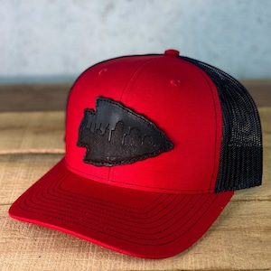 May include: A red and black trucker hat with a black leather patch featuring a city skyline in the shape of an arrowhead.
