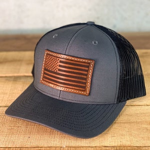 May include: A gray and black trucker hat with a brown leather American flag patch on the front.