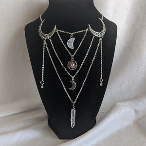 May include: A silver layered necklace with multiple crescent moon and star charms, a sun charm, and a clear quartz crystal pendant.