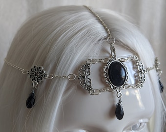 Op de afbeelding: Een zilveren ketting hoofdband met een zwarte ovale edelsteen en hangende zwarte druppelvormige kristallen.