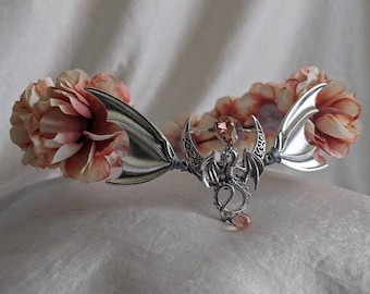 May include: A silver dragon with a pink crystal pendant, attached to a silver crescent moon, is surrounded by two large, pale pink fabric flowers. The flowers are attached to the dragon with silver wire.