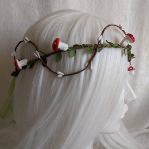 May include: A brown twig crown with white and red mushroom accents. The crown is decorated with small white flowers and green leaves.