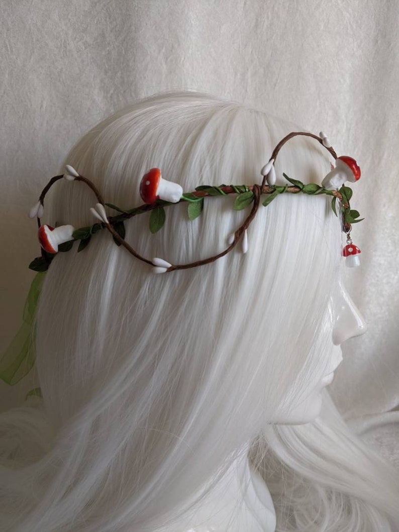 May include: A brown twig crown with white and red mushroom accents. The crown is decorated with small white flowers and green leaves.