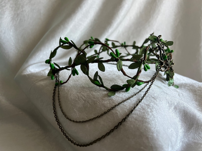 May include: A green leaf crown with a chain detail. The crown is made of faux leaves and berries and has a delicate chain attached to the back.