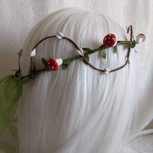 May include: A brown twig crown with white cotton buds and red and white mushroom accents. The crown is decorated with green leaves.