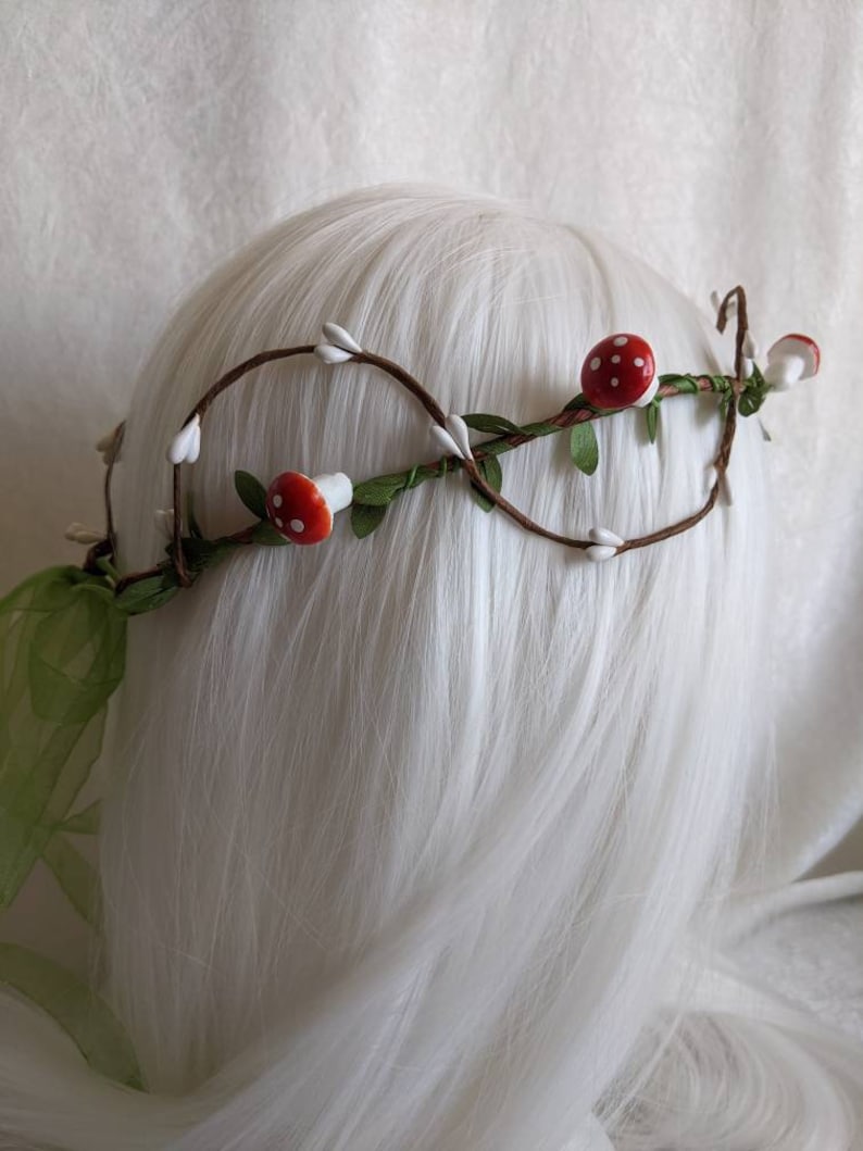 May include: A brown twig crown with white cotton buds and red and white mushroom accents. The crown is decorated with green leaves.