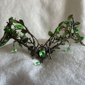 May include: A green and bronze crown with a large green gemstone in the center. The crown is made of wire and has a leafy design.