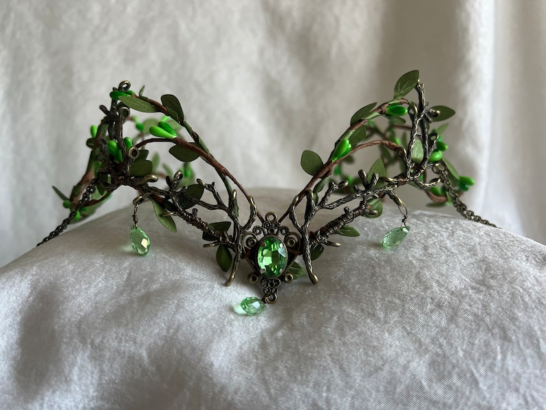 May include: A green and bronze crown with a large green gemstone in the center. The crown is made of wire and has a leafy design.