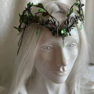 May include: A green and bronze crown with a large green gemstone in the center. The crown is made of branches and leaves, and has a chain hanging from the side.