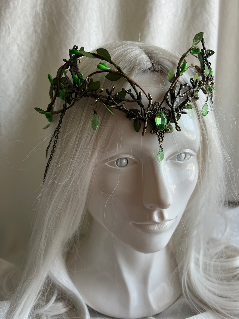 May include: A green and bronze crown with a large green gemstone in the center. The crown is made of branches and leaves, and has a chain hanging from the side.
