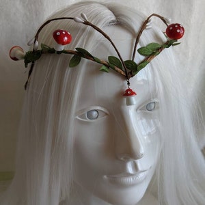 May include: A white headband with brown branches, green leaves, and red and white mushroom accents.