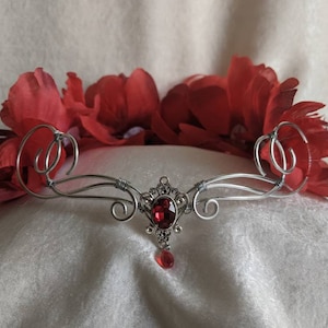 Peut inclure: Une couronne en fil d'argent ornée d'une pierre rouge centrale et d'une gemme en forme de goutte pendante. Des fleurs en tissu rouge complètent le design. Idéale pour les occasions spéciales.