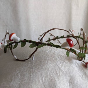 May include: A white and brown twig crown with red and white mushroom accents. The crown is decorated with small white flowers and green leaves.