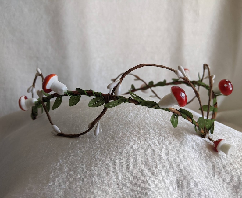 May include: A white and brown twig crown with red and white mushroom accents. The crown is decorated with small white flowers and green leaves.