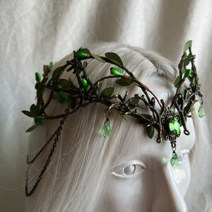 May include: A green leaf and twig crown with a large green gemstone in the center. The crown is made of metal and has a chain on each side.