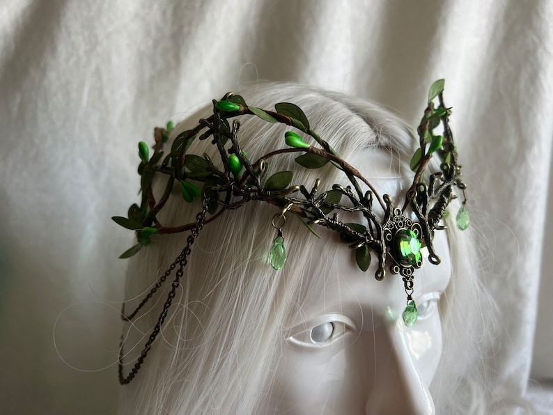 May include: A green leaf and twig crown with a large green gemstone in the center. The crown is made of metal and has a chain on each side.