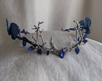 Op de afbeelding: Een zilveren kroon met blauwe bloemen van stof en een halve maan ontwerp. De maan heeft een blauwe edelsteen in het midden.