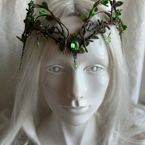 May include: A crown made of metal branches with green leaves and green gem accents. The crown is on a white mannequin head with long white hair.
