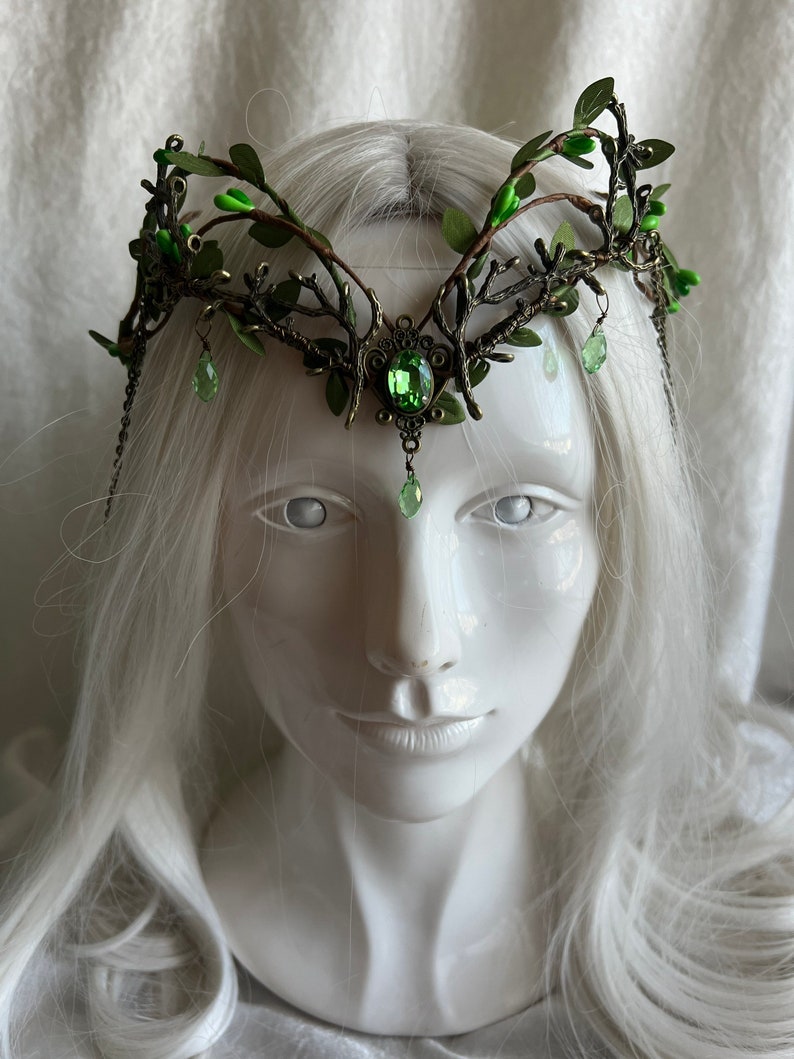 May include: A crown made of metal branches with green leaves and green gem accents. The crown is on a white mannequin head with long white hair.