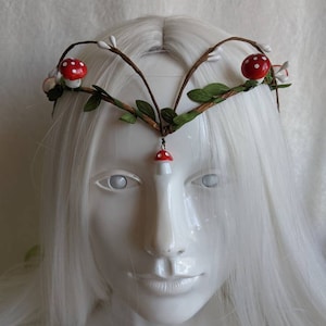 May include: A white headband with brown branches, green leaves, and red and white mushroom accents.