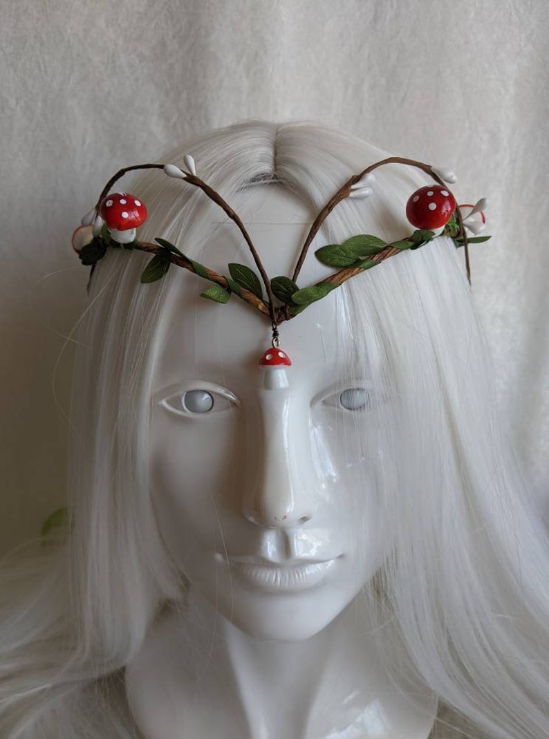 May include: A white headband with brown branches, green leaves, and red and white mushroom accents.