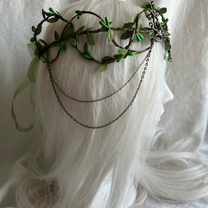 May include: A green leaf crown with brown branches and metal chains. The crown is on a white mannequin head.