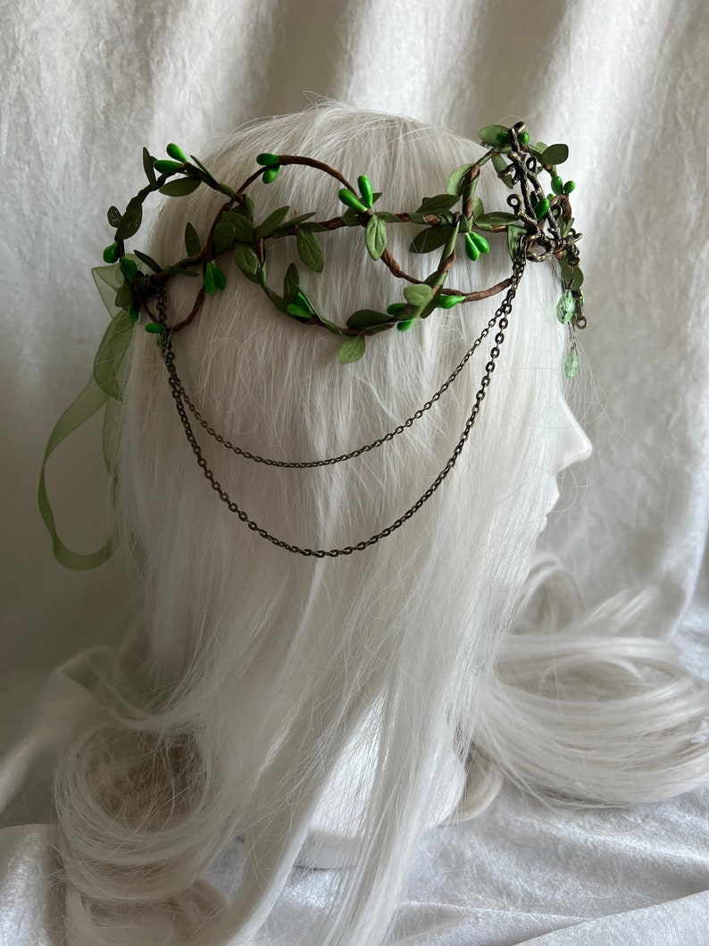 May include: A green leaf crown with brown branches and metal chains. The crown is on a white mannequin head.