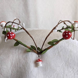 May include: A handmade twig crown with red and white mushroom accents. The crown is decorated with small white flowers and green leaves.