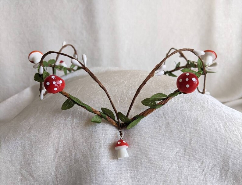 May include: A handmade twig crown with red and white mushroom accents. The crown is decorated with small white flowers and green leaves.
