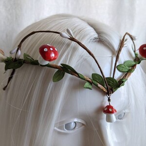 May include: A whimsical forest-themed headband with brown twigs, green leaves, and red and white mushroom accents.